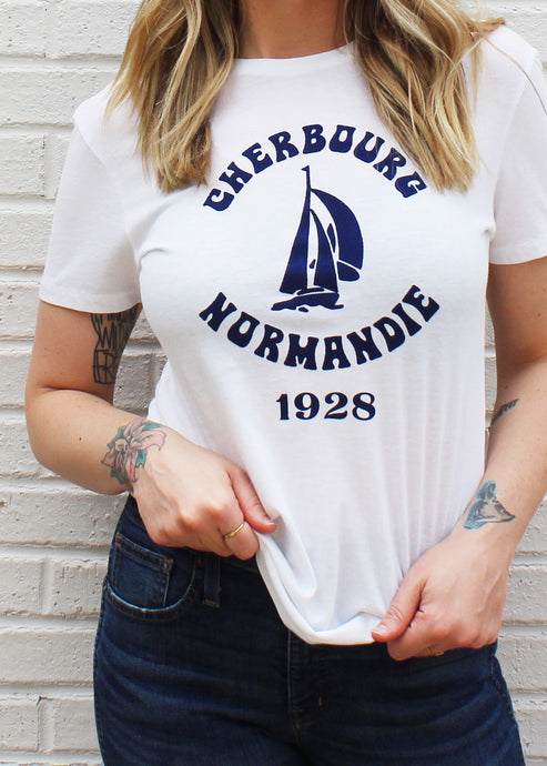 Female modeling white unisex graphic tee with navy print that reads Cherbourg Normandie 1928 around a sailboat.